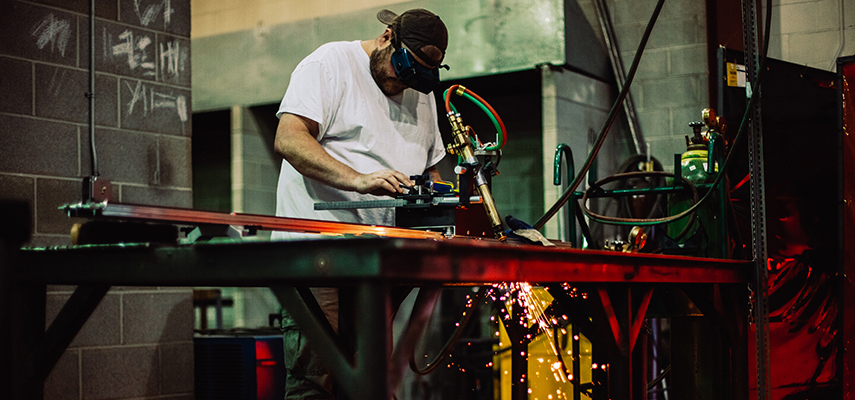 A student wearing a welding mask fabricating and/or manufacturing a piece of technology on a welder's table.
